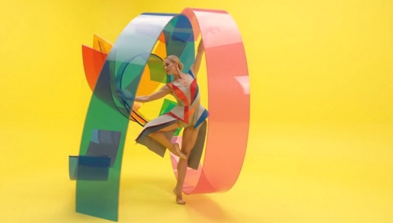 Dancer alongside a monumental multicolored translucent ribbon structure on yellow.